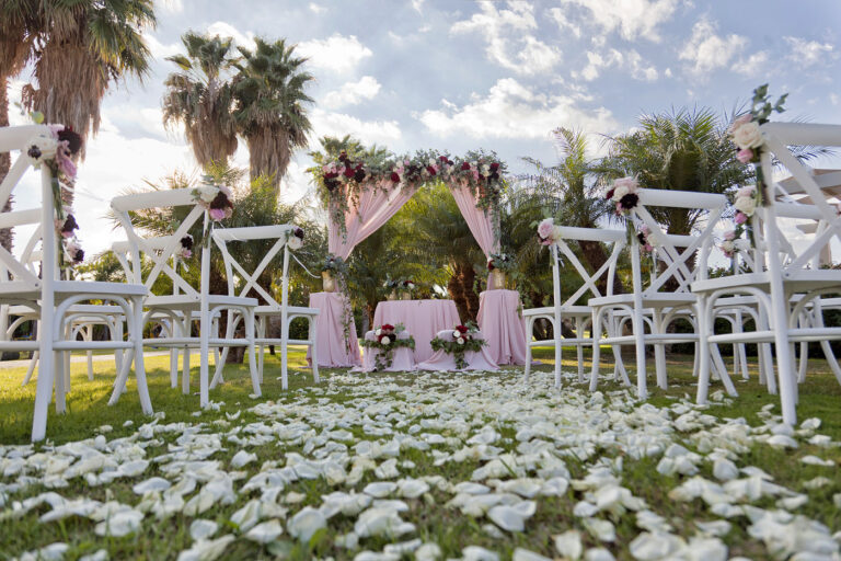 Garden ceremony among palms, with a blush flowered arch and a carpet of white petals, Oggi Sposi & Events
