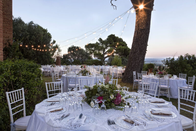 Round tables with low flower arrangements in a garden under string lights at dusk, Oggi Sposi & Events