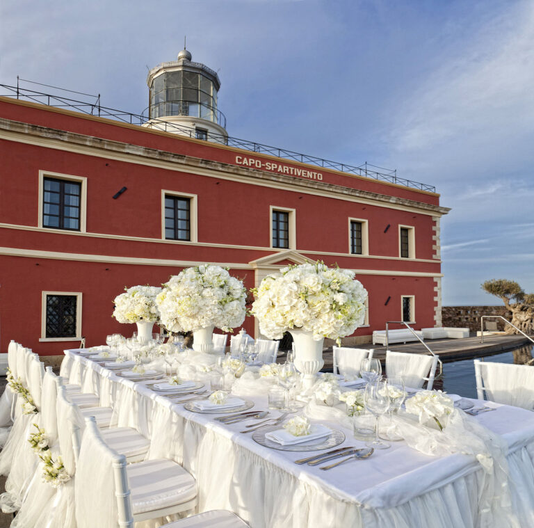 Long table dressed in white with tall white arrangements at the Capo Spartivento lighthouse, Oggi Sposi & Events
