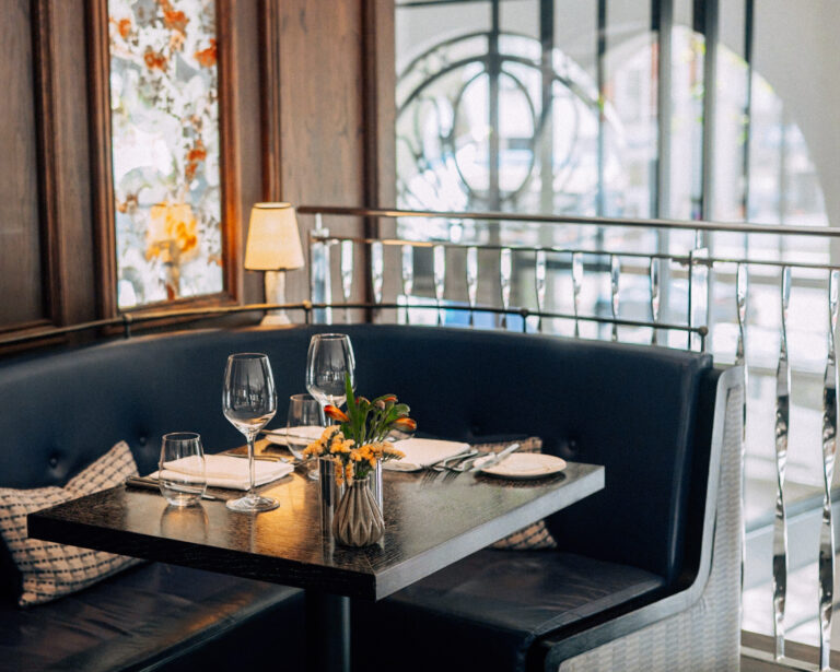 A banquette table in Indigo beside the arched windows, laid for lunch, One Aldwych