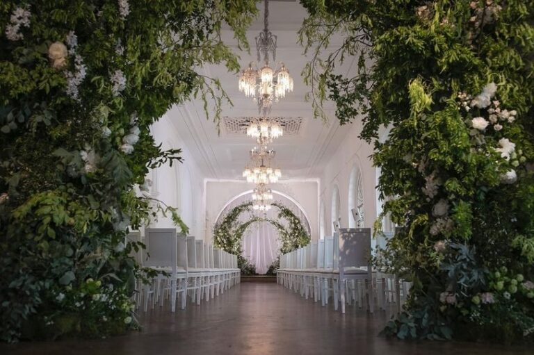 The Gallery dressed with a lush green-and-white floral arch and twin chandeliers for a ceremony, One Marylebone