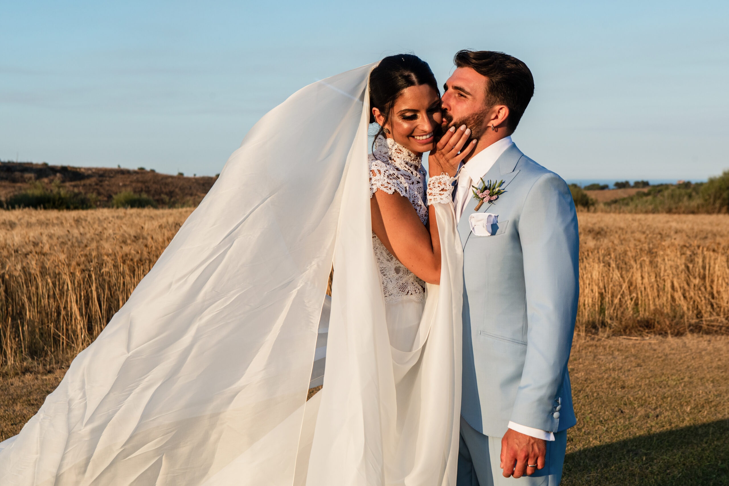 The couple in a cut wheat field at dusk, her veil blown wide across the frame, Pangiunìa Wedding