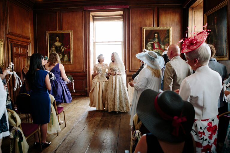 Two brides walk through their guests in a panelled room hung with portraits, Paola De Paola Photography