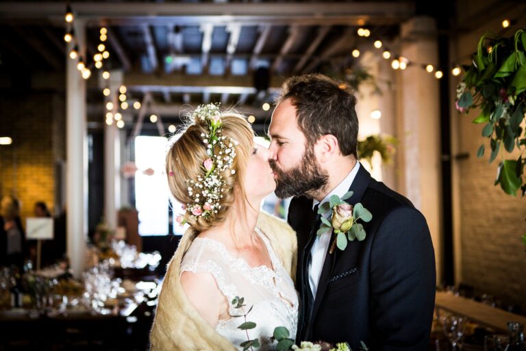 A bride with a flower crown kisses her groom under strings of lights, Paola De Paola Photography