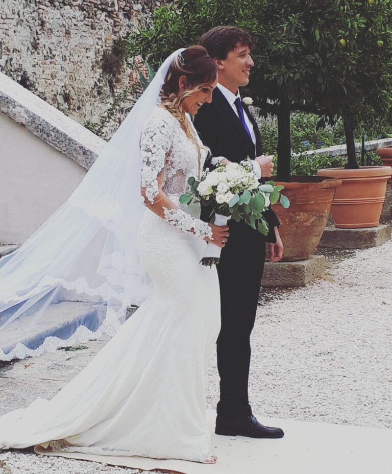 Bride in a long veil and lace sleeves standing beside the groom on a garden stair, Paola Giancola Makeup