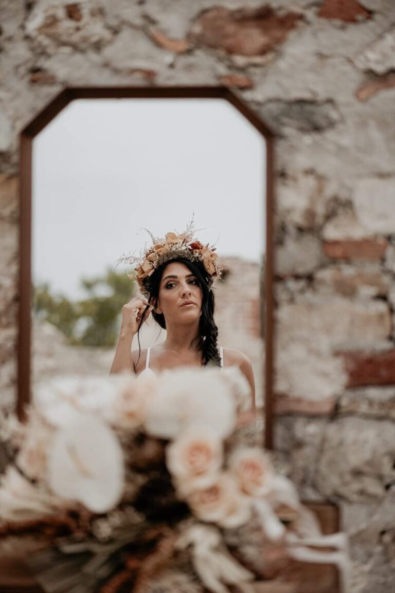 Bride with a flower crown reflected in an arched mirror, with a bouquet in the foreground, Paola Giancola Makeup