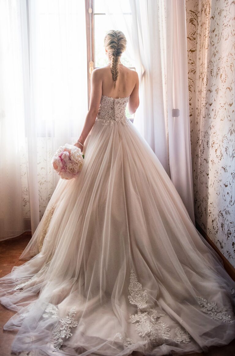 Bride seen from behind in a tulle gown at a tall window, with a braided hairstyle, Paola Giancola Makeup