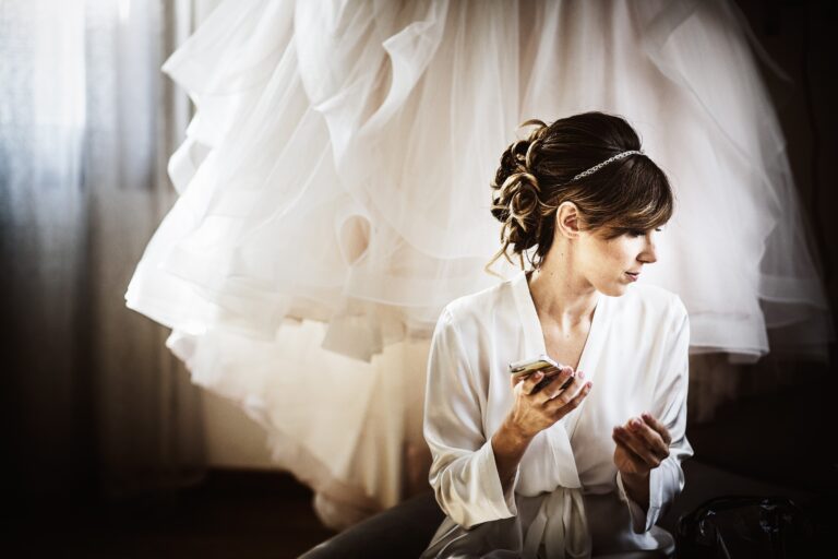 Bride with an updo and headband seated in front of her skirts, holding a phone, Paola Giancola Makeup