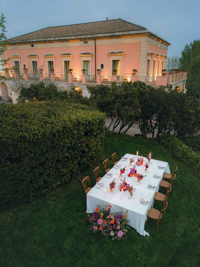A table dressed in bright flowers on the lawn below a pink villa, Giardini di Villa Fago, Sicily, Paola Lapi Luxury Event