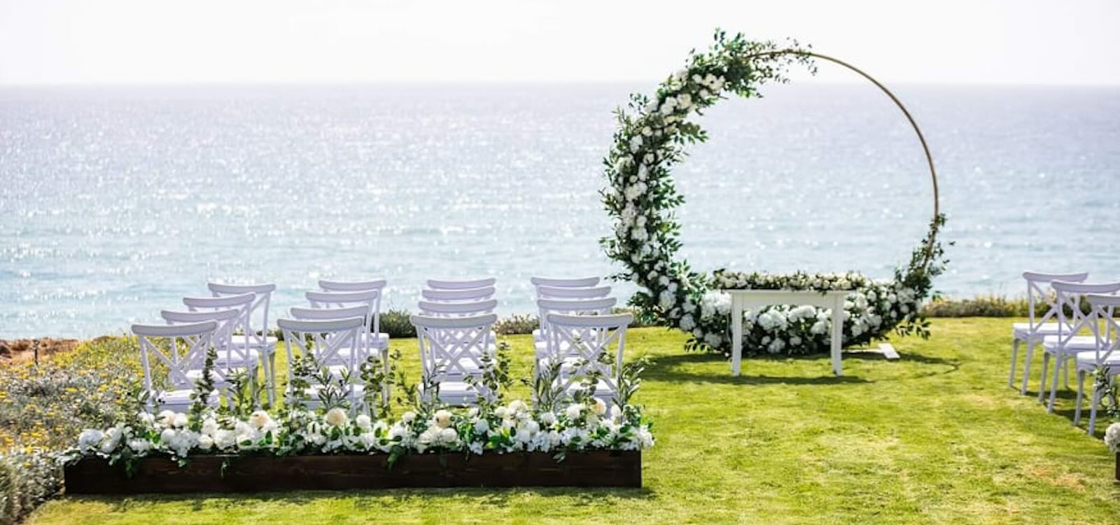 Circular floral arch and white chairs for a ceremony on a seafront lawn, Alassos, Paphos Weddings Made Easy
