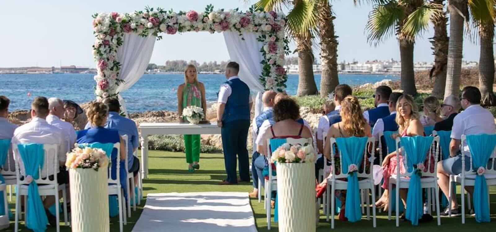 Guests seated for a ceremony under a floral canopy with turquoise chair sashes, Olympic Lagoon Resort, Paphos Weddings Made Easy