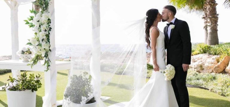 The couple kiss beneath a white floral arch with the veil lifting, Olympic Lagoon Resort, Paphos Weddings Made Easy