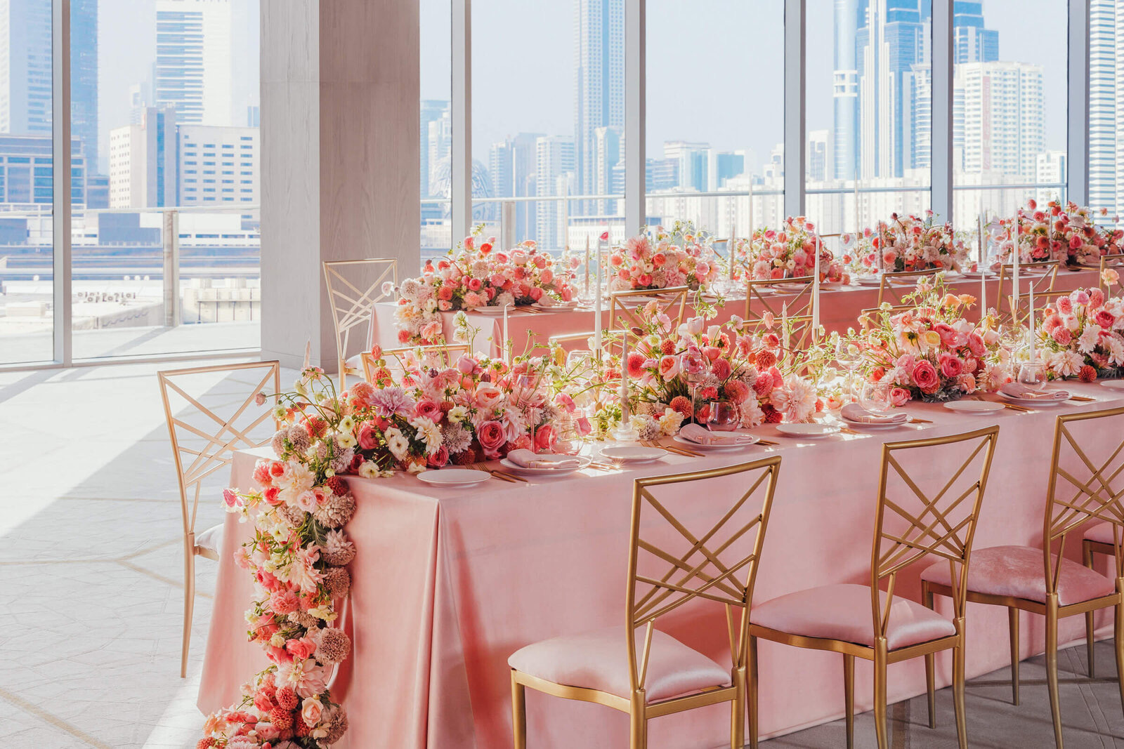 Long pink-clothed table with floral runner in a glass-walled ballroom above a city skyline, Patrizia Cilli Events Italy