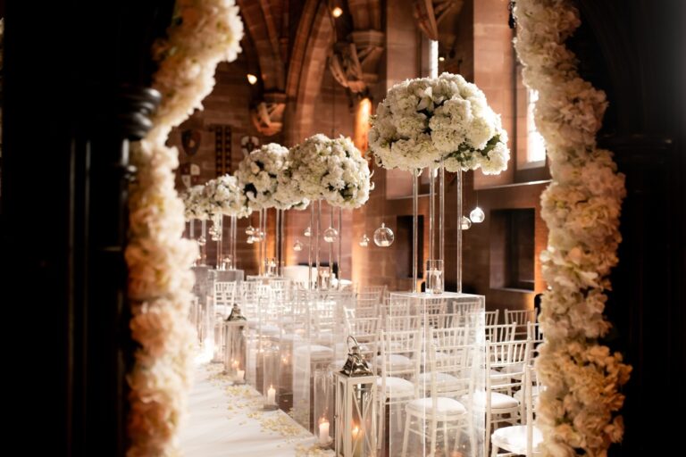 The Great Hall dressed for a ceremony, tall white floral arrangements lining the candlelit aisle beneath stone arches, Peckforton Castle