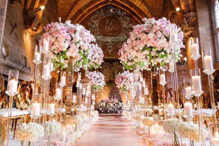 The Great Hall aisle framed by towering pink and white floral pillars and candles, a monogrammed backdrop beyond, Peckforton Castle