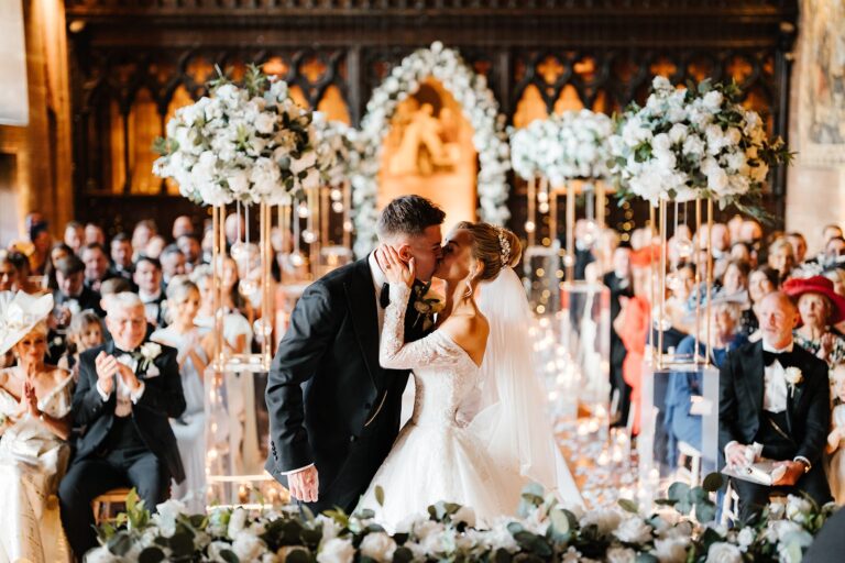 A couple's first kiss at the altar in the Great Hall, guests applauding among white florals, Peckforton Castle