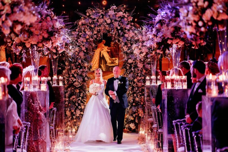 A bride walks the candlelit aisle on her father's arm between blush floral arches, Peckforton Castle