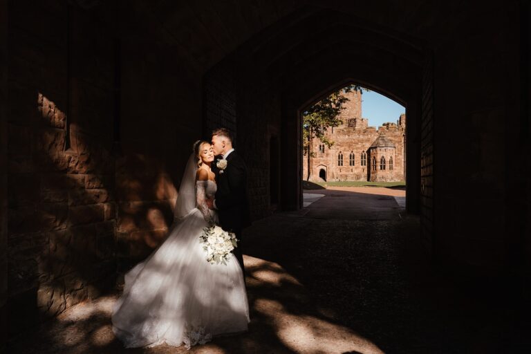 A couple in the shadowed stone gatehouse archway, the sunlit courtyard beyond, Peckforton Castle