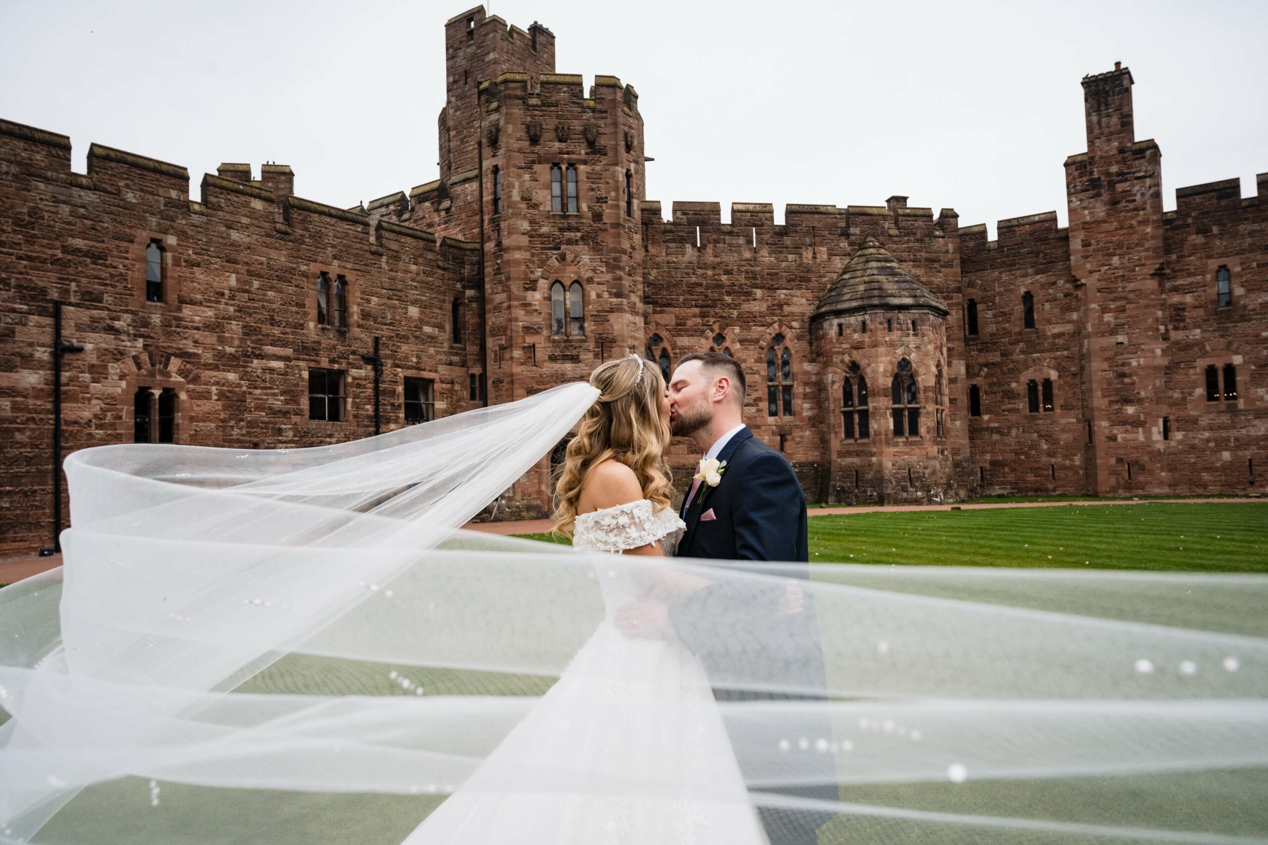 A couple kiss in the castle courtyard, the bride's veil sweeping across the frame, red sandstone towers behind, Peckforton Castle