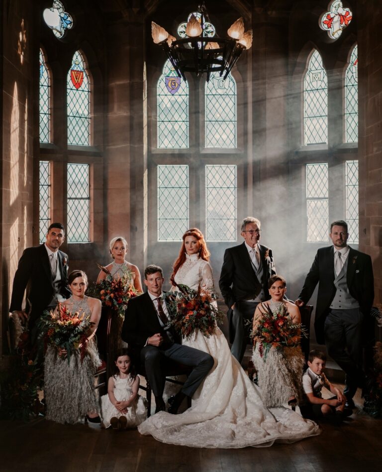 A wedding party portrait beneath the castle's heraldic stained-glass windows, with autumn bouquets, Peckforton Castle