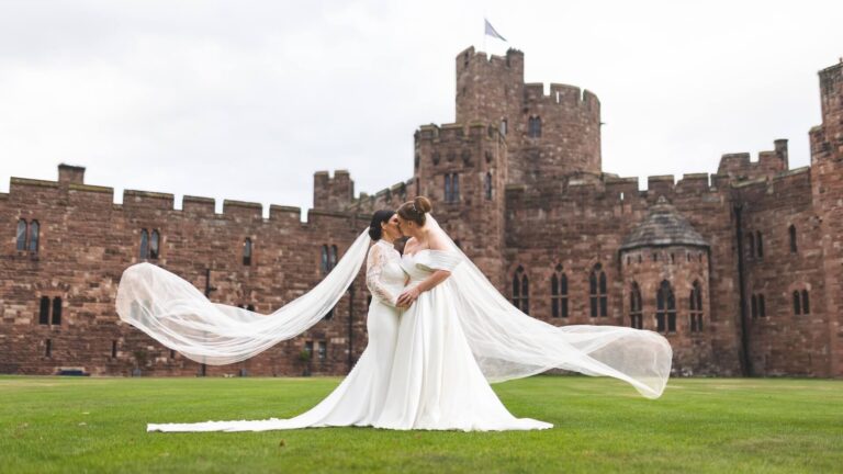 Two brides embrace on the lawn, veils sweeping, the red sandstone castle behind them, Peckforton Castle