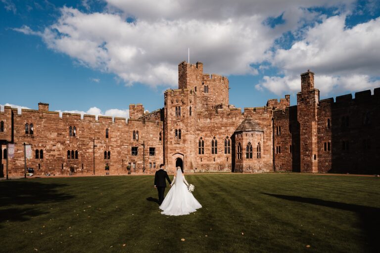 A couple walk across the lawn toward the castle under a bright Cheshire sky, Peckforton Castle