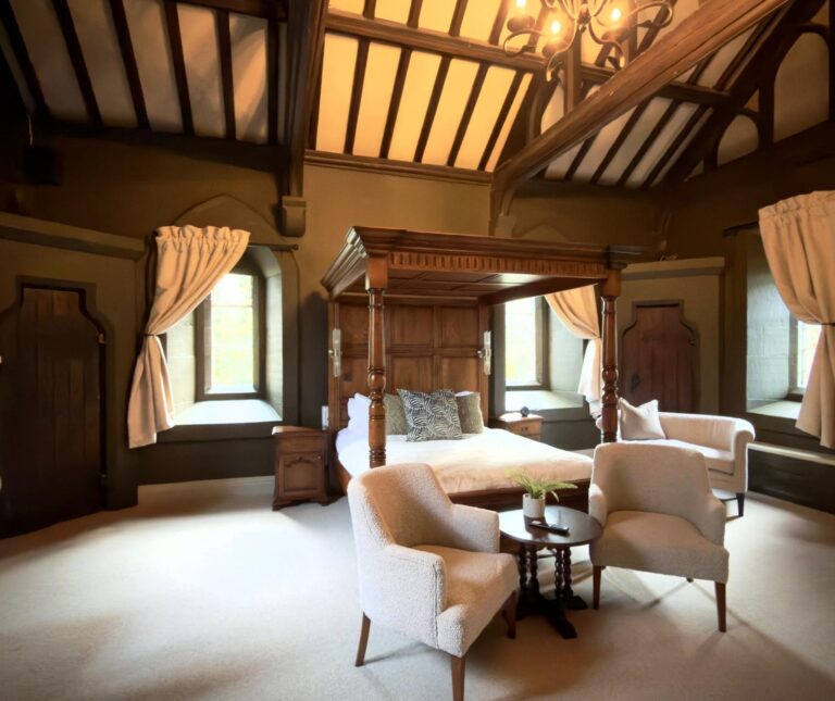 A grand suite beneath a beamed and vaulted ceiling, a carved four-poster and cream armchairs by mullioned windows, Peckforton Castle