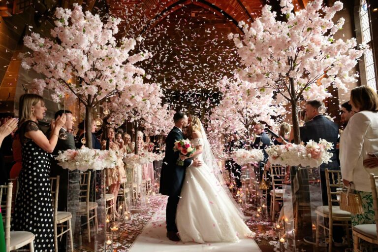 A couple beneath blossom trees in a shower of petals, guests seated either side, Peckforton Castle