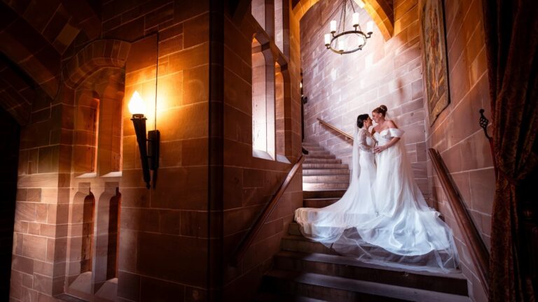 Two brides on the grand sandstone staircase beneath a wall lantern, Peckforton Castle