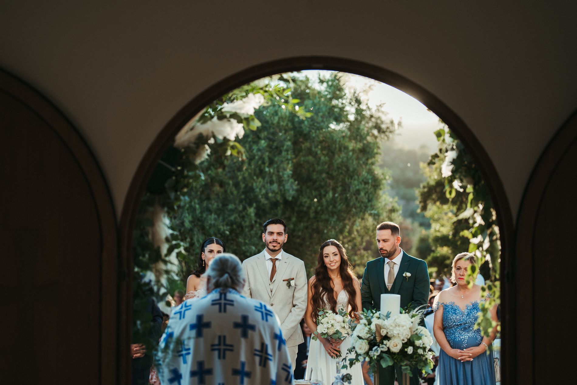 The couple and priest framed through a stone archway during the ceremony, Photomad