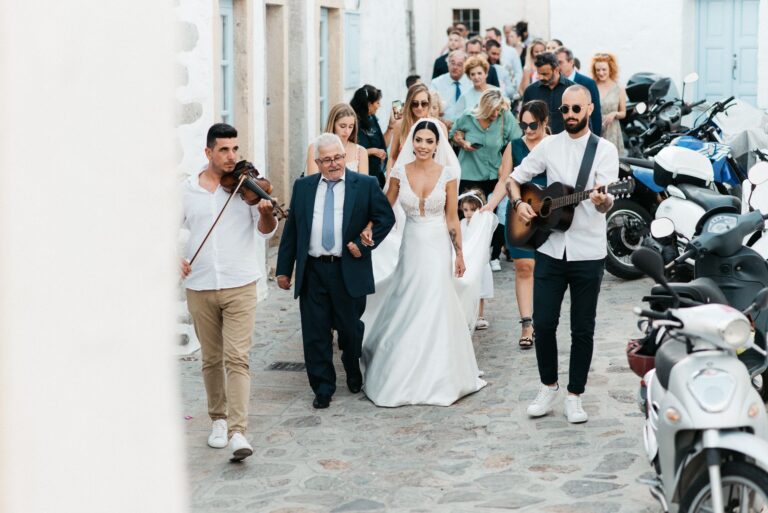 The bride walks a whitewashed lane with a violinist and guitarist playing beside her, Photomad