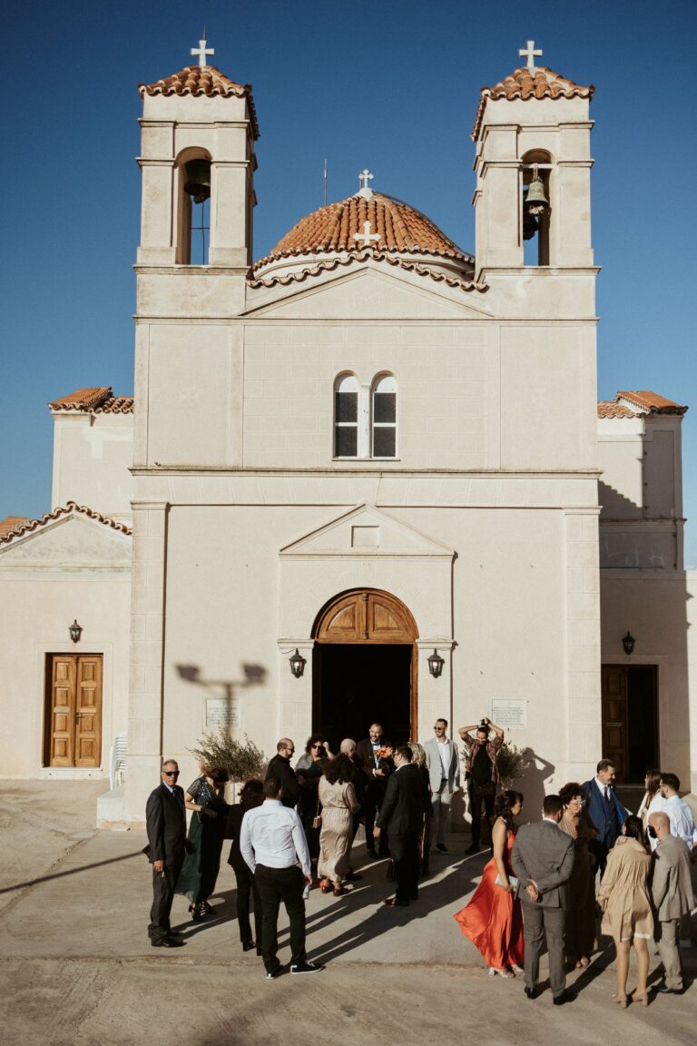 Guests gather outside a white twin bell tower church, Photomad