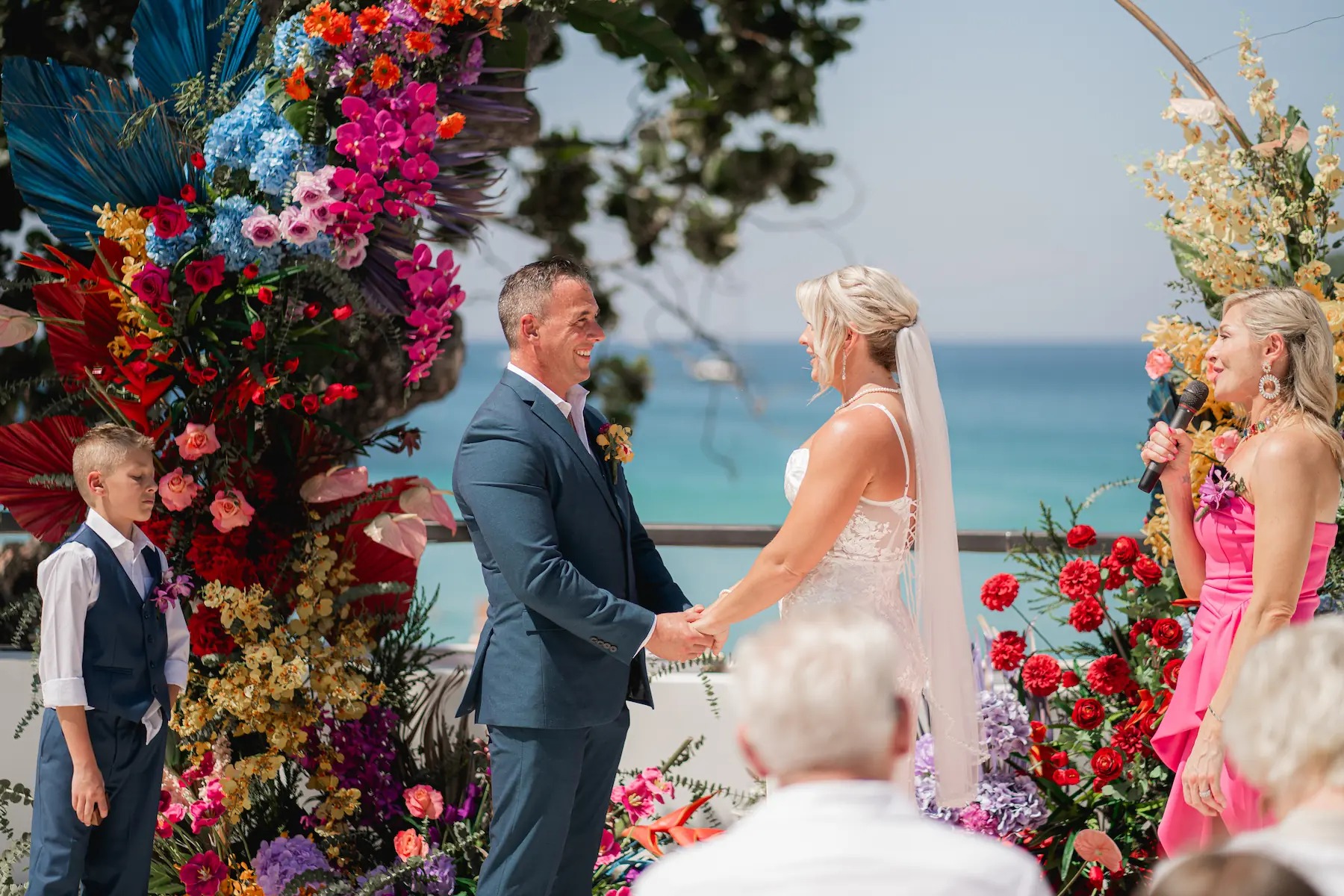 A couple exchange vows beneath a tall arch of bright tropical flowers on a terrace above the sea at The Boathouse Phuket, Phuket Romance