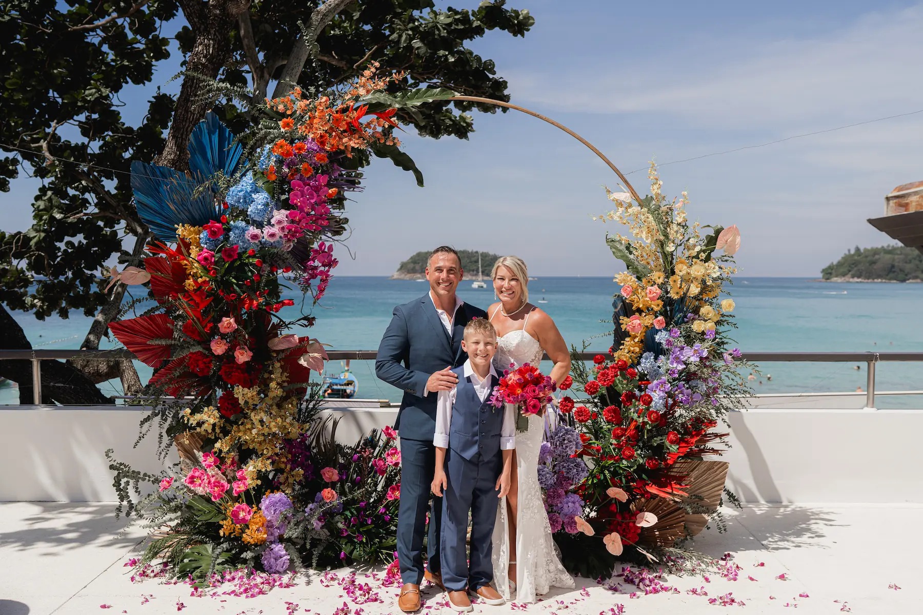 A couple and their young son stand in front of a colourful floral arch on a white terrace, with the bay and a small island behind, Phuket Romance
