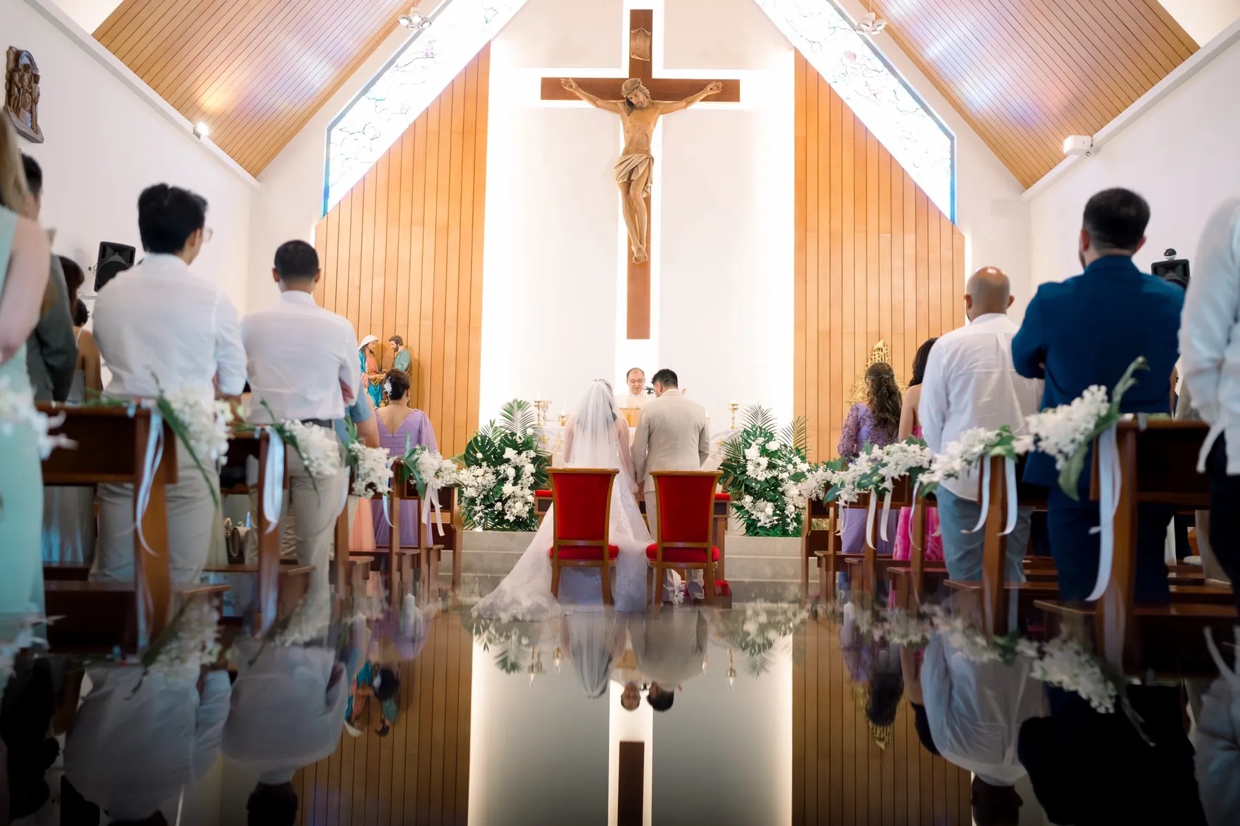 A wedding mass at the white chapel of St. Joseph Church, Phuket, with a wooden crucifix over the altar and flowers along the pews, Phuket Romance