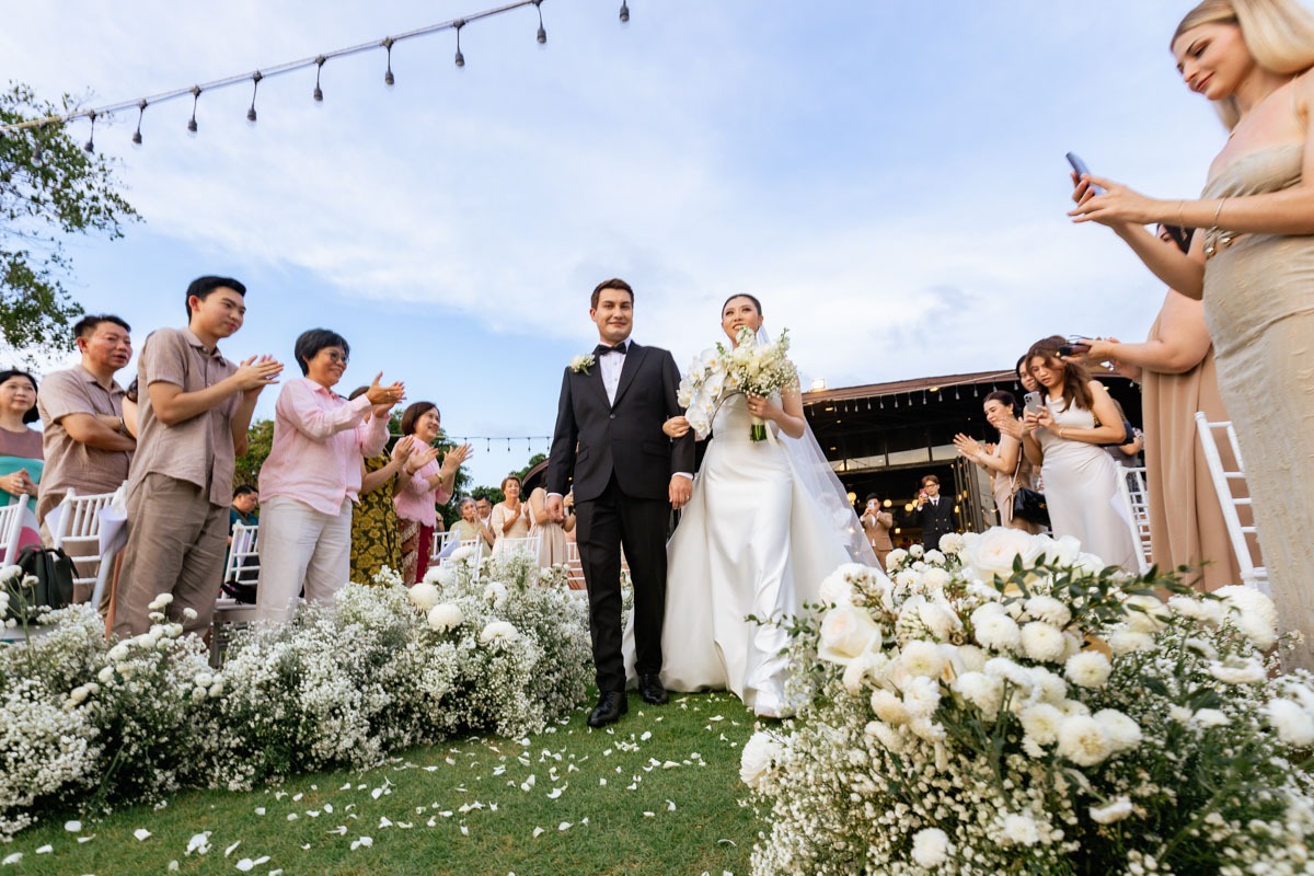 A bride and groom walk a lawn aisle edged with white roses and gypsophila as guests applaud at Le Méridien Khao Lak, Phuket Romance
