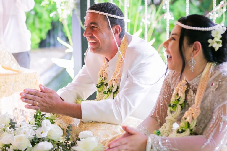 A couple in traditional Thai wedding dress with ceremonial headbands and garlands laugh during their ceremony at The Pavilions Phuket, Phuket Romance