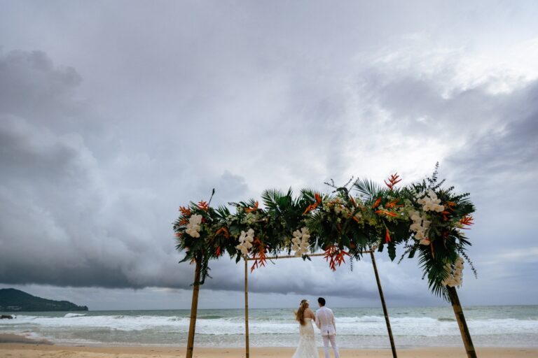 A couple at a beach ceremony arch of palm leaves and orange flowers under a dramatic cloudy sky at The Surin Phuket, Phuket Romance