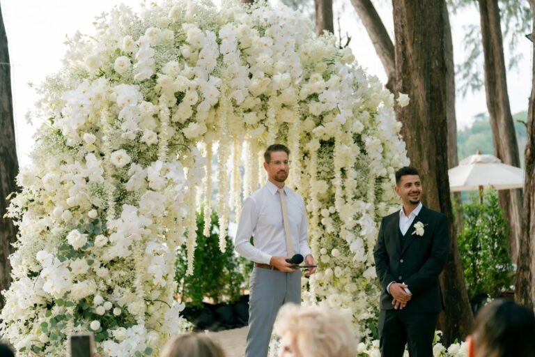 A dense arch of white flowers and hanging blooms behind the groom and celebrant at a beachside ceremony at InterContinental Phuket, Phuket Romance
