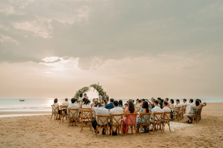 Guests seated in a horseshoe of wooden chairs on the sand at sunset around a small floral arch, Devasom Khao Lak, Phuket Romance