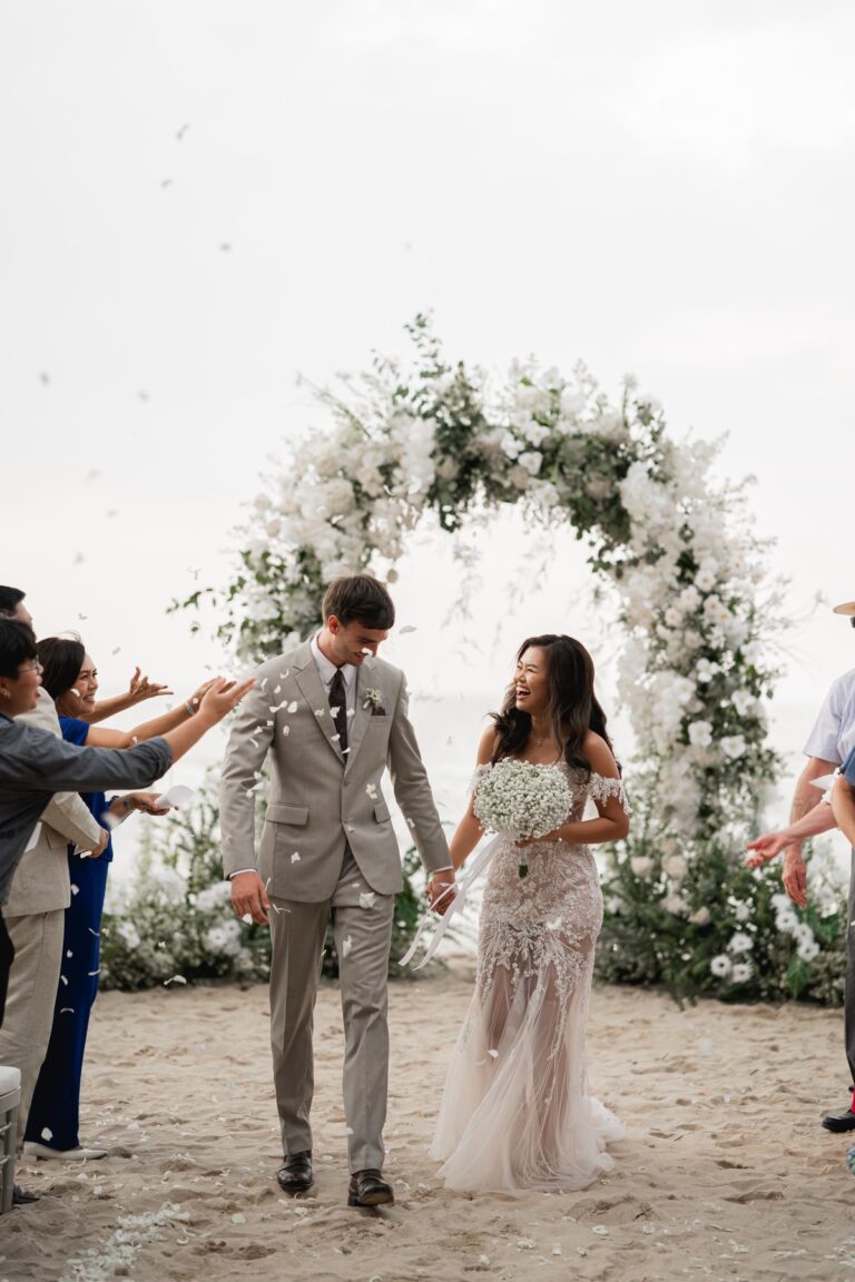 A couple walk hand in hand through a white flower arch on the sand as guests throw petals, Avani Khao Lak, Phuket Romance