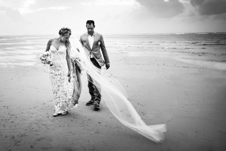 A bride and groom walk along the shore in black and white, the bride's long veil trailing in the wind, at The Slate Phuket, Phuket Romance
