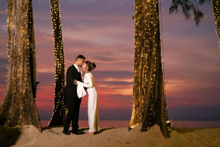 A couple kiss on the beach at dusk between two trees wound with fairy lights under a pink and purple sky, Phuket Romance
