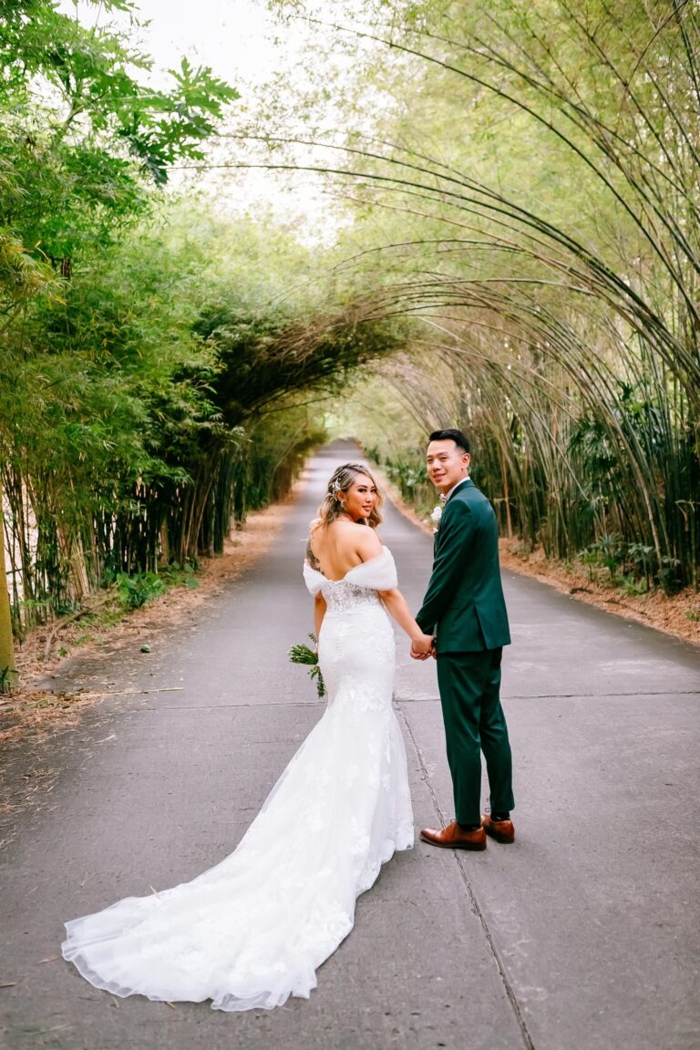 A bride in a lace-trained gown and a groom in a green suit stand on a bamboo-lined road at The Pavilions Phuket, Phuket Romance