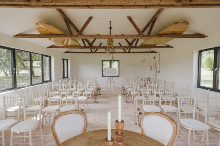 The Boathouse set for a ceremony, rows of white chairs under exposed timber trusses and paddleboards stored in the roof, Pinkmead Estate