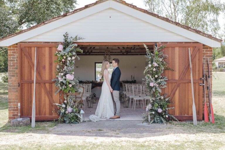 A couple framed by the open timber doors of the Boathouse, dressed with tall floral arrangements, with tables laid inside, Pinkmead Estate