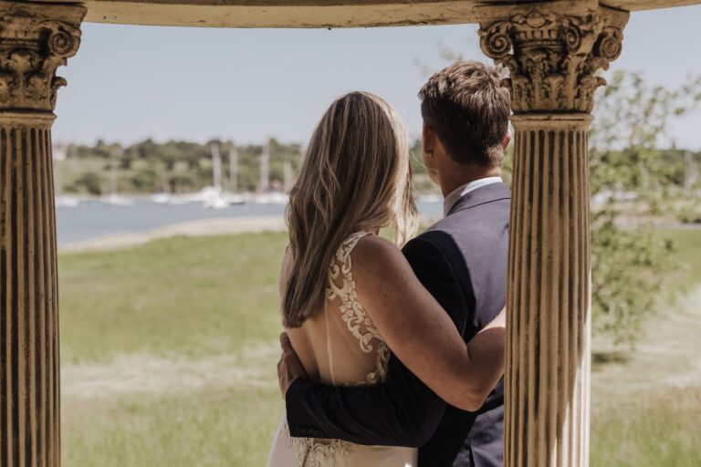 A couple seen from behind between the temple's fluted columns, looking out over the river and moored yachts, Pinkmead Estate