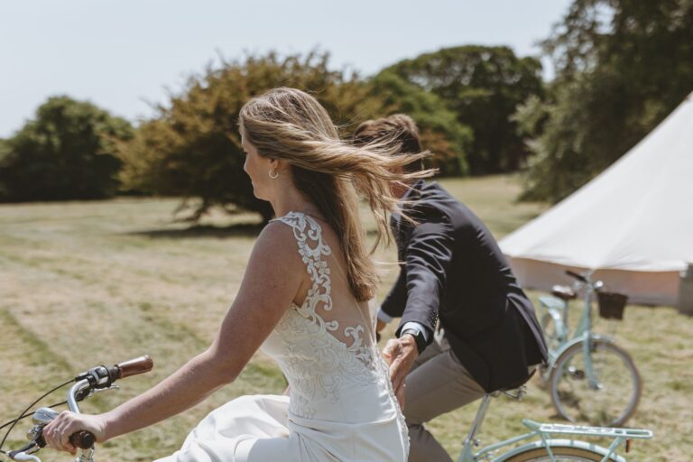 A bride and groom riding the estate's bicycles across the lawn past a bell tent, Pinkmead Estate