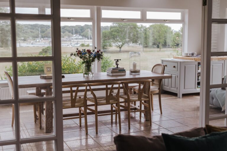 The dining table in the main house, with wishbone chairs, garden flowers and a wide window over the river, Pinkmead Estate