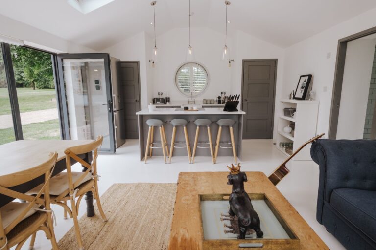 An open-plan living space with a kitchen island, porthole window, sofa and dining table, and doors open to the garden, Pinkmead Estate